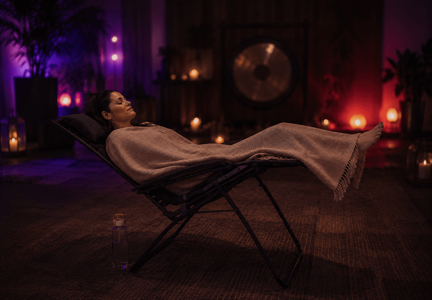 Side-profile of a person fully reclined in a zero-gravity chair under a soft blanket, candles and a glowing gong in the warmly lit background