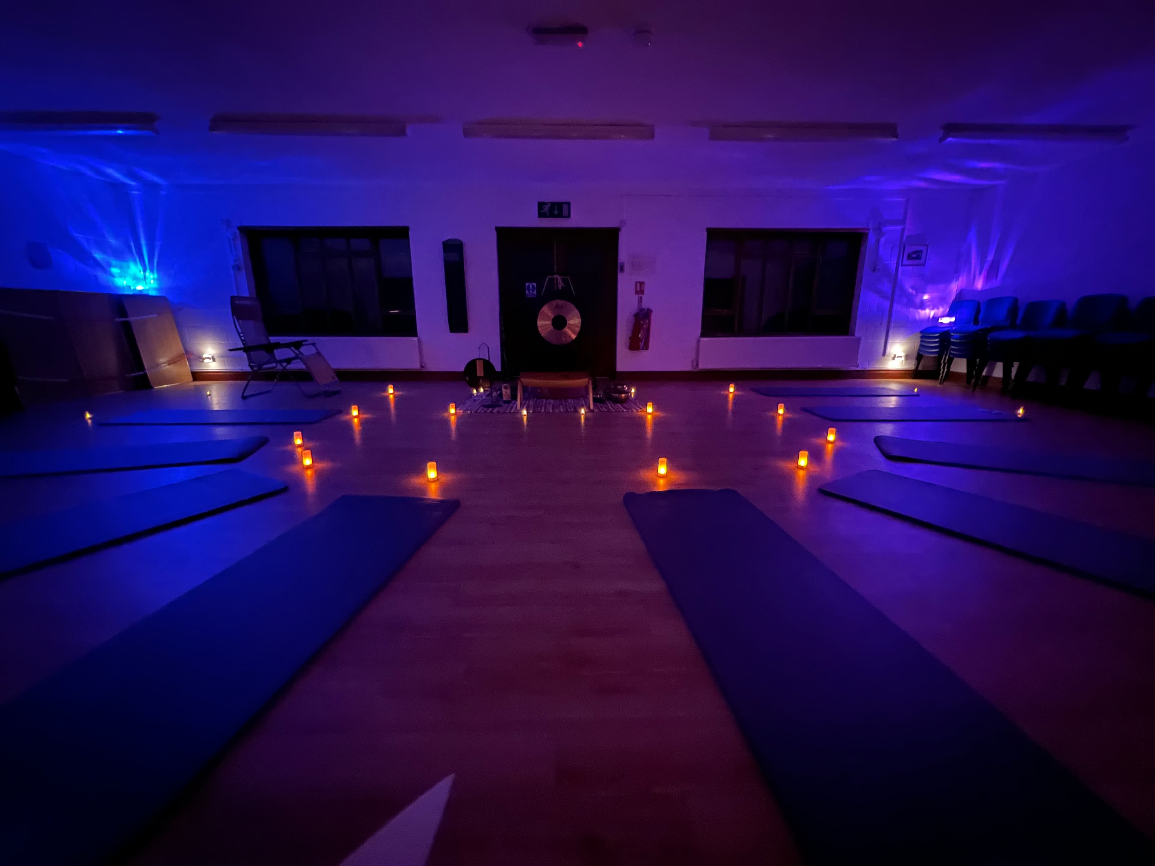 Sound bath room set up with yoga mats radiating from a centre gong, candles glowing between them under soft blue and purple light