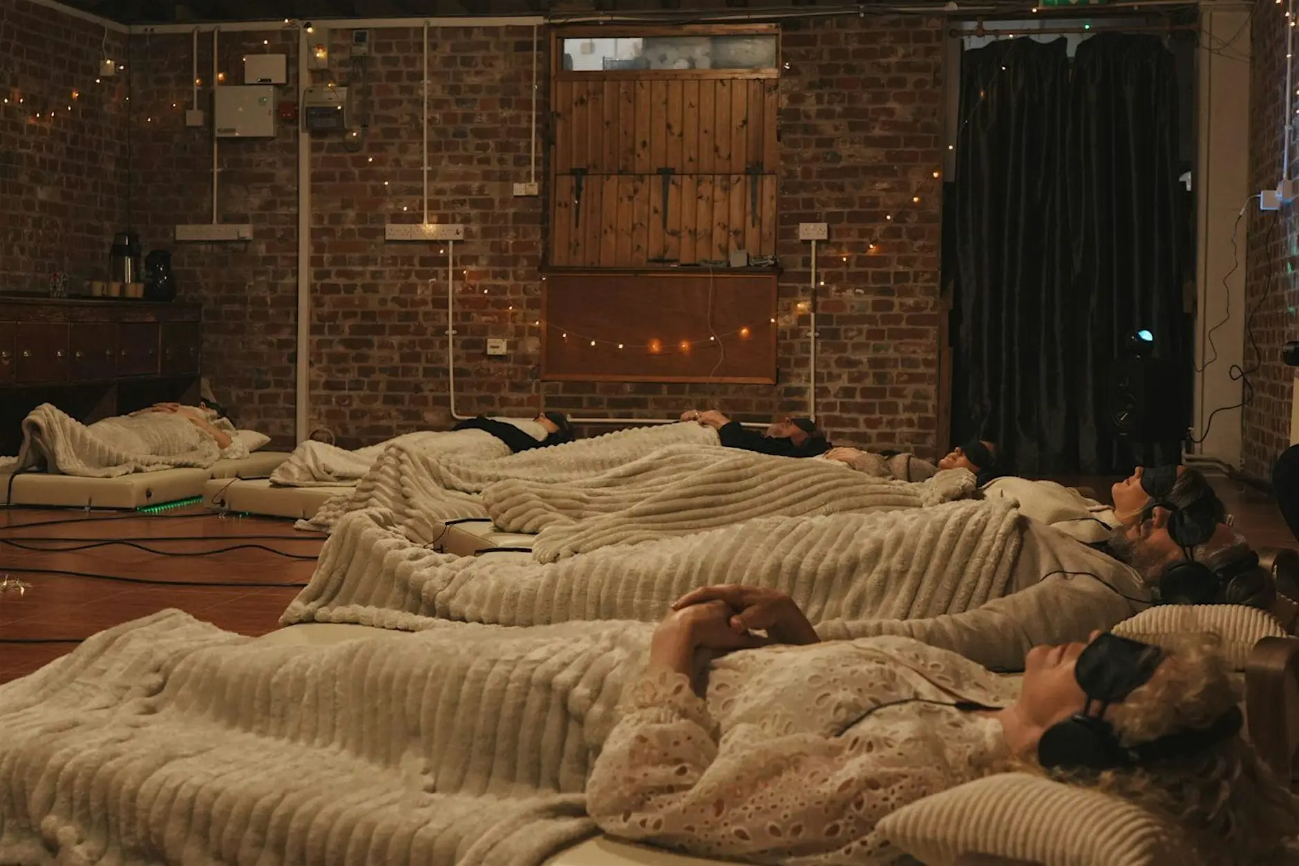 Participants resting on Onizion vibroacoustic therapy beds wearing eye masks and closed-back headphones in a candlelit brick room